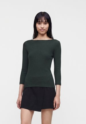 Femme aux cheveux noirs raides portant un haut à manches longues vert foncé et une jupe courte noire, debout devant un fond gris clair.