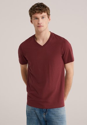 Jonge man met krullend haar, gekleed in een effen bordeauxrode V-hals T-shirt en een blauw spijkerbroek, staand tegen een licht beige achtergrond.