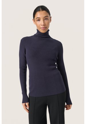 Marinblå ribbestrikket turtleneck sweater med lange ærmer, tætsiddende design, blød tekstur og flad nederkant. Båret med sorte skræddersyede bukser.