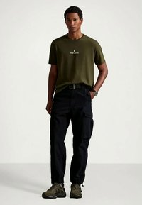 Man standing wearing olive green Ralph Lauren t-shirt, black cargo pants, black belt, olive green sneakers, and wristwatch against plain background.