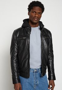 Schwarze Lederjacke mit abnehmbarer Kapuze, ausgestattet mit Reißverschlusstaschen und einer figurbetonten Passform, getragen über einem hellblauen Hemd und Jeans.