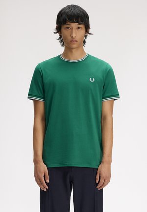 Giovane uomo in piedi, rivolto in avanti, che indossa una maglietta verde a maniche corte con bordino bianco e pantaloni scuri su uno sfondo semplice.