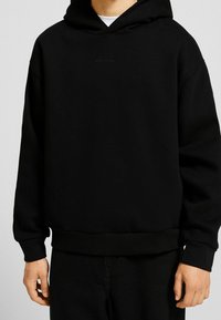 Svart huvtröja gjord av mjukt tyg, med en kenguruficka och minimalistisk logotypdetalj på bröstet. Ribbad mudd och nederkant.