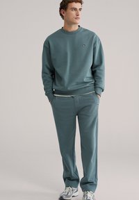 Tirkizna majica dugih rukava s rebrastim manšetama i rubom, uparena s odgovarajućim hlačama. Na oba odjevna predmeta nalazi se mali izšivani logotip.