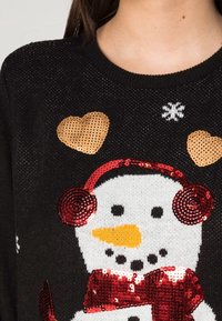 Suéter negro de punto con un diseño de muñeco de nieve que presenta auriculares de lentejuelas rojas, una nariz de zanahoria naranja y acentos dorados en forma de corazón.
