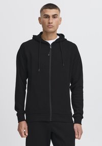 Sort zip-up hættetrøje med hætte, tekstureret stof, forlommer og kontrasterende syninger. Har justerbare snore og ribbede manchetter.