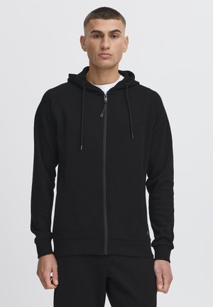 Zwarte rits-hoody met een capuchon, gestructureerde stof, voorzakken en contrasterende naden. Voorzien van verstelbare trekkoorden en geribbelde manchetten.