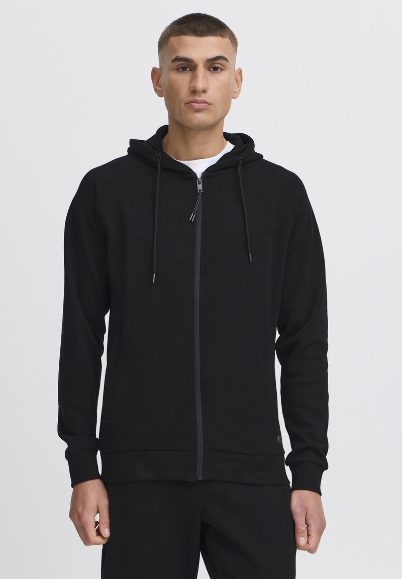 Sort zip-up hættetrøje med hætte, tekstureret stof, forlommer og kontrasterende syninger. Har justerbare snore og ribbede manchetter.