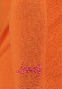 Oranges Pullover mit geripptem Textur und einer pinkfarbenen gestickten "Lovely"-Details am Ärmel. Einfaches Design, glatte Stoffoberfläche.
