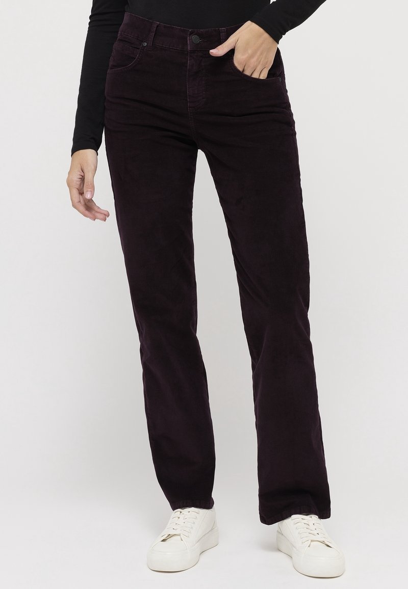 Femme portant un pantalon en velours côtelé violet foncé avec des baskets blanches, une main dans la poche, debout devant un fond clair uni.