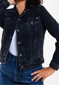 Musta farkkutakki istuvalla mallilla, jossa hopeiset napit, kaksi rintataskua ja teksturoitu kangas. Yhdistetty sinisiin farkkuihin.