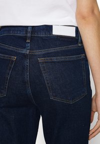 Mörkblå denimjeans med medelhög midja, fem fickor och kontrasterande orange sömmar. Vit etikett på baksidan av midjebandet.