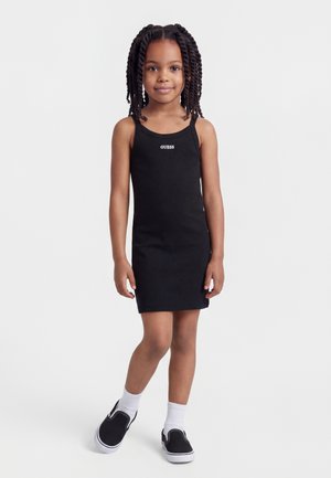 Ragazza giovane con capelli intrecciati che indossa un vestito nero senza maniche con logo "GUESS", calze bianche e scarpe slip-on nere, in piedi davanti a uno sfondo bianco.