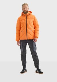 Chaqueta de plumas naranja con capucha, con un acolchado en forma de rombos, cremalleras negras y dos bolsillos laterales. Combinada con pantalones cargo oscuros y zapatillas negras.