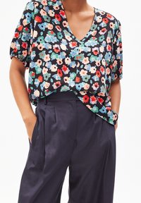 Persona que lleva una blusa corta negra con estampado floral metida dentro de pantalones sueltos de talle alto en color marino, con las manos en los bolsillos.