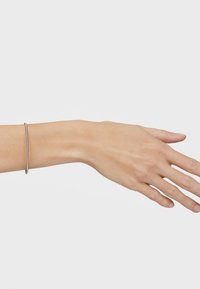 Brazalete delgado y plateado en una mano, con una superficie lisa y pulida. El brazalete es circular y descansa sobre la muñeca, contra la piel desnuda.