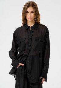 Black sheer button-up shirt with two chest pockets, long sleeves, and a relaxed fit, displaying a lightweight, translucent fabric.