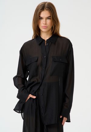 Camisa negra de botón transparente con dos bolsillos en el pecho, mangas largas y un corte relajado, exhibiendo una tela ligera y translúcida.