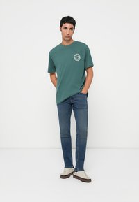 Camiseta de algodón verde con un logo circular blanco en el lado izquierdo del pecho, combinada con pantalones vaqueros azul denim y zapatillas de color claro.