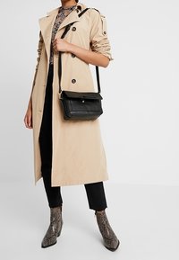 Beige Trenchcoat mit hochgerollten Ärmeln, schwarzer Umhängetasche, schwarzen Hosen und grauen Schlangenleder-Stiefeletten.