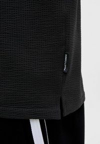 Chemise noire en textile texturé à manches courtes avec un motif à carreaux, dotée d'une fente sur le côté et d'une petite étiquette indiquant "PROJECT CORE."