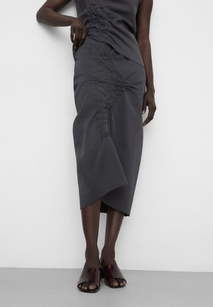 Jupe midi gris foncé texturée avec ourlet asymétrique, portée avec des sandales slides ouvertes à l'avant de couleur foncée, sur une personne debout contre un mur blanc.