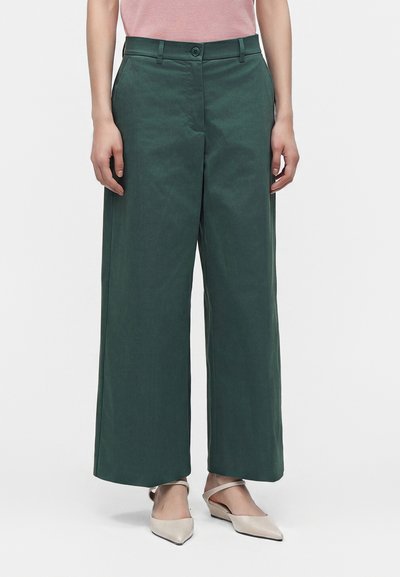Femme portant un pantalon large vert foncé, un haut rose pâle et des ballerines beiges à bouts pointus avec des brides à la cheville, debout sur fond blanc.