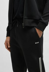 Schwarze Jogginghose mit Kordelzug am Bund und seitlichen Taschen, mit einem weißen "BOSS"-Logo und weißen Streifen an den Seiten.