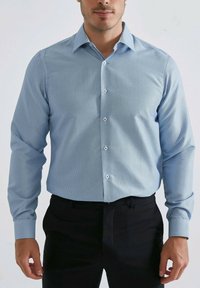 Chemise à manches longues bleu clair avec un motif géométrique, col boutonné et boutons blancs, associée à un pantalon noir.