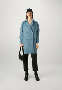 Woman wearing a blue double-breasted coat, black cropped pants, black boots, black cap, and holding a black handbag against a plain background.