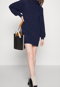 Chunky stickad marinblå klänning med hög krage, långa ärmar och en lös passform, i kombination med en svart tote-väska och ankelskor.
