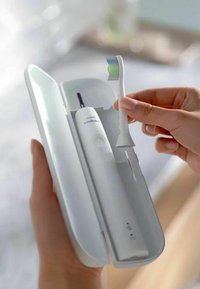 Spazzolino elettrico in un case bianco. Lo spazzolino è bianco con una testina di setole verde e blu. Il case ha un design elegante e curvo.