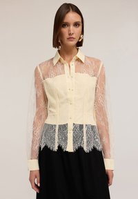Blouse van kant en chiffon met een gele lijfje, doorschijnende lange mouwen en ingewikkelde kantdetails aan de zoom, aangevuld met een knoopsluiting.