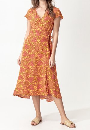 Frau trägt ein Wickelkleid mit gelbem und rotem Muster, kurzen Ärmeln und beige gewebte flache Sandalen, steht vor einem einfarbigen Hintergrund.