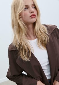Bruine buitenjas met een brede kraag en een relaxte pasvorm, gedragen over een witte ronde hals shirt. Lange, golvende blonde haren. Zachte, gladde textuur.