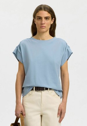 Femme aux longs cheveux bruns portant une chemise ample bleu clair à manches courtes, un pantalon beige et tenant une lanière de sac marron.