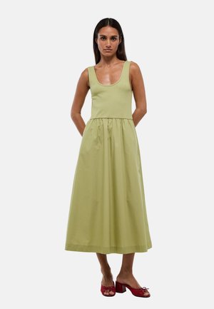 Femme portant une robe midi sans manches vert clair et des chaussures rouges à talons ouvertes, debout les mains dans le dos sur fond blanc uni.