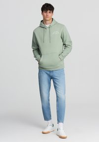 Felpa verde chiaro con cordini, tasca frontale e scritte bianche. Abbinata a jeans azzurri e sneaker bianche, indossata con calze di lunghezza media.