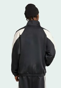Corta-vento atlético preto com painéis de destaque brancos, gola alta e faixa elástica na cintura. Tecido texturizado com um corte solto. Boné preto usado.