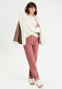 Pantalons en velours côtelé rose, pull crème et manteau doublé en shearling marron. Baskets blanches et roses avec une semelle texturée. Modèle posé contre un fond uni.
