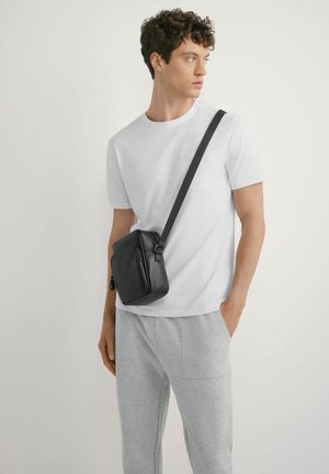 Młody mężczyzna w białej koszulce i szarych spodniach, niosący czarną torbę na ramię z szerokim paskiem, patrzący na bok.