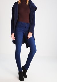 Kvinna klädd i en marinblå kappa över en brun polo, mörkblå tighta jeans och svarta ankelstövlar, stående med det ena benet något korsat.