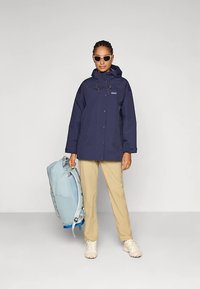 Μπλε ναυτικό αδιάβροχο μπουφάν με κουκούλα και κορδόνι, συνδυασμένο με ανοιχτό καφέ παντελόνι. Το άτομο κρατάει μια γαλάζια τσάντα πλάτης και φοράει κρεμ αθλητικά παπούτσια.