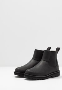 Botas de tornozelo em couro preto com painéis laterais elásticos, bico arredondado e sola de borracha texturizada. Apresenta alças para facilitar o calçar.