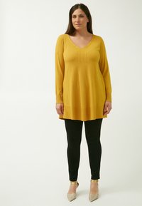 Tunic gialla senape con scollatura a V e cuciture verticali testurizzate. Abbinata a leggings neri e tacchi a punta metallici. Tessuto a maglia liscia.