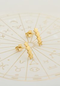 Guldstjärnformade örhängen med en polerad yta och en unik design, utställda på en dekorativ astrologisk bakgrund.
