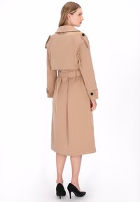 Beiger Trenchcoat mit tailliertem Gürtel, Schulterklappen, Knopfdetails und mittellanger Länge bis zur Mitte der Wade. Kombiniert mit schwarzen High-Heels.