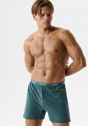 Mężczyzna bez koszulki patrzący w dół, ubrany w zielone bokserki z nazwą marki "ATLANTIC" na gumce, z rękami założonymi za plecami.