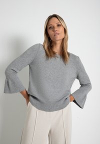 Hellgrauer Strickpullover mit ausladenden gerippten Ärmeln und einem gerippten Halsausschnitt. Präsentiert mit lockerer Passform und über cremefarbenen Hosen getragen.