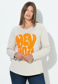 Ljusbeige långärmad skjorta med en stor orange texturerad "HEY YOU!" grafik på framsidan, ihop med mörkblåjeans.